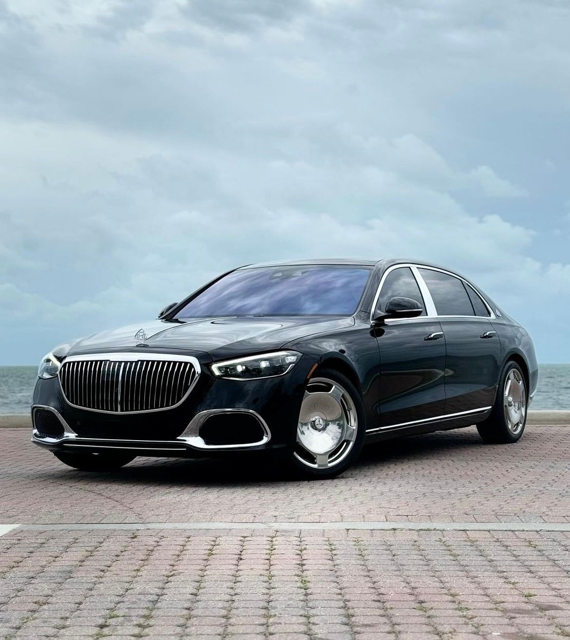 Luxury black sedan parked on a paved area near the ocean with a cloudy sky in the background.